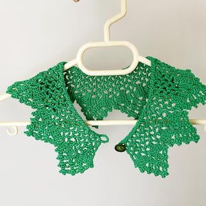 May include: Green crocheted collar with a button closure. The collar is hanging on a white hanger.