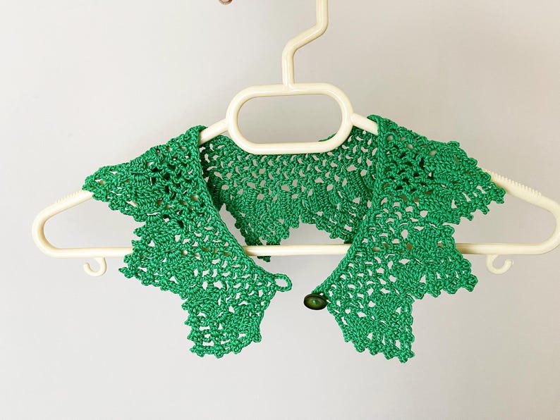 May include: Green crocheted collar with a button closure. The collar is hanging on a white hanger.