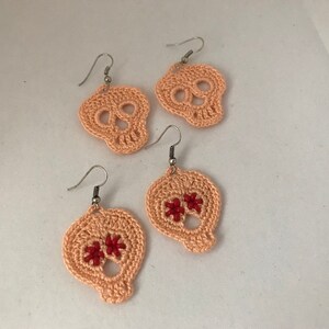 May include: Three pairs of crocheted skull earrings. Two pairs are peach colored and one pair has red flower accents.