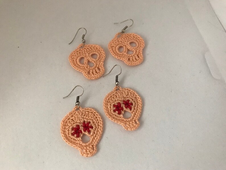 May include: Three pairs of crocheted skull earrings. Two pairs are peach colored and one pair has red flower accents.