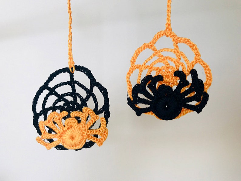 May include: Two crocheted spider web ornaments with spiders hanging from the center. One web is black with an orange spider, and the other is orange with a black spider.