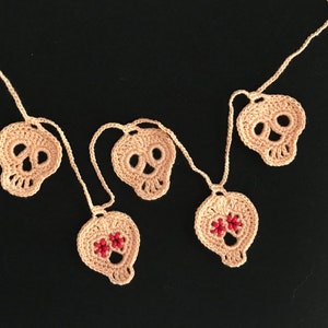 May include: A garland of five crocheted skull ornaments, strung together with white yarn. The skulls are beige and have black eyes and noses. Two of the skulls have red bead accents.