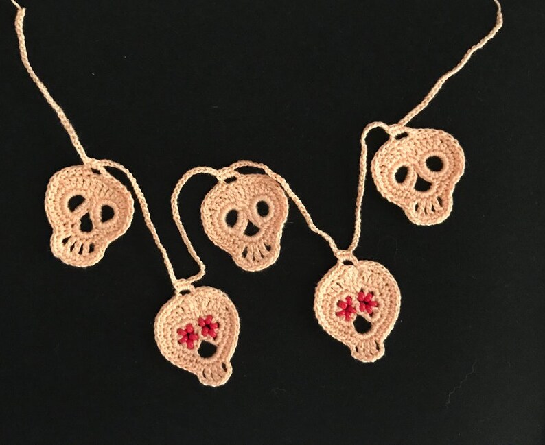 May include: A garland of five crocheted skull ornaments, strung together with white yarn. The skulls are beige and have black eyes and noses. Two of the skulls have red bead accents.
