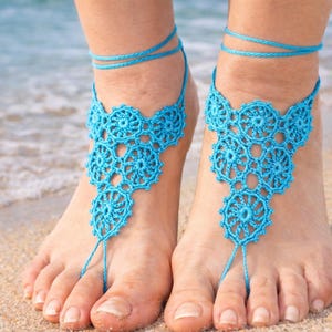 Puede incluir: Sandalias descalzas de ganchillo turquesa. Presentan un diseño floral y están unidas por un hilo que se enrolla alrededor del tobillo y el dedo del pie. Las sandalias se muestran en una playa de arena con el mar de fondo.