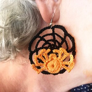 May include: A pair of black and orange crocheted earrings featuring a spider design. The earrings are round and have a web-like pattern.