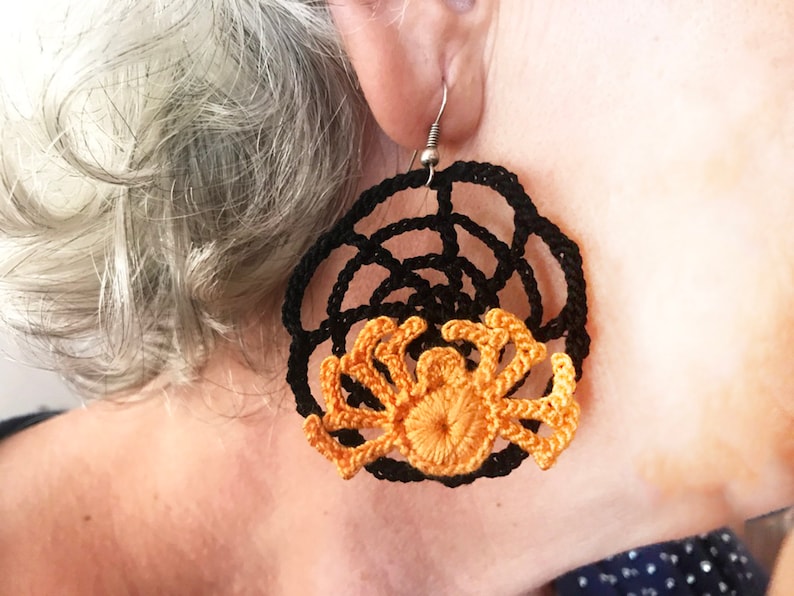 May include: A pair of black and orange crocheted earrings featuring a spider design. The earrings are round and have a web-like pattern.