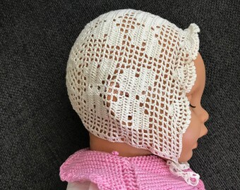 Puede incluir: Un gorro de bebé blanco de ganchillo con un borde festoneado. El gorro está hecho con un delicado patrón de calado.