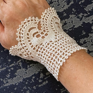 Könnte beinhalten: Ein weißes Häkelarmband mit einem zarten Spitzenmuster. Das Armband ist aus feinem Garn gefertigt und hat einen zarten, femininen Look.