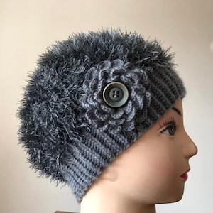May include: Grey knitted beanie with a fuzzy pom pom and a crocheted flower detail. The flower has a large button in the centre.