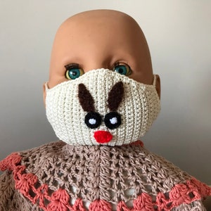 Puede incluir: Una m&aacute;scara facial de crochet blanca con un dise&ntilde;o de reno marr&oacute;n. La m&aacute;scara tiene ojos negros y una nariz roja.
