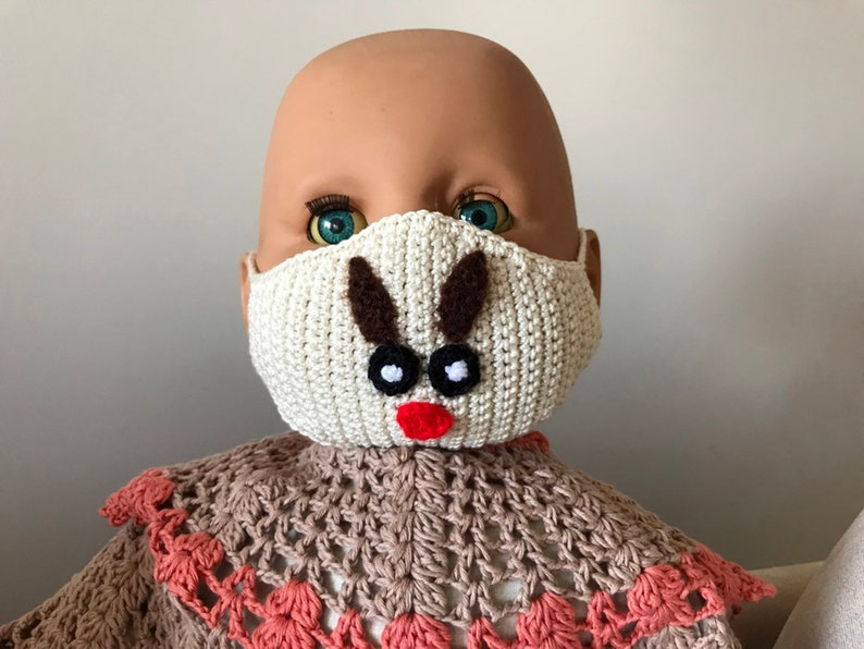 Puede incluir: Una m&aacute;scara facial de crochet blanca con un dise&ntilde;o de reno marr&oacute;n. La m&aacute;scara tiene ojos negros y una nariz roja.