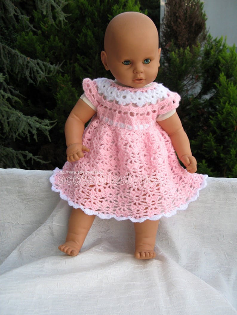 May include: A pink crocheted dress with white trim, worn by a doll with blue eyes. The dress has a scalloped hem and a white crocheted collar.