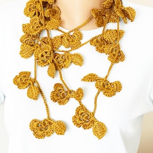 Pode incluir: Um colar longo de crochê com um design floral. O colar é feito de fio amarelo mostarda e apresenta delicadas flores e folhas de crochê.