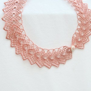 May include: A delicate pink crocheted necklace with a white bead accent. The necklace is made of intricate lacework and has a scalloped edge.