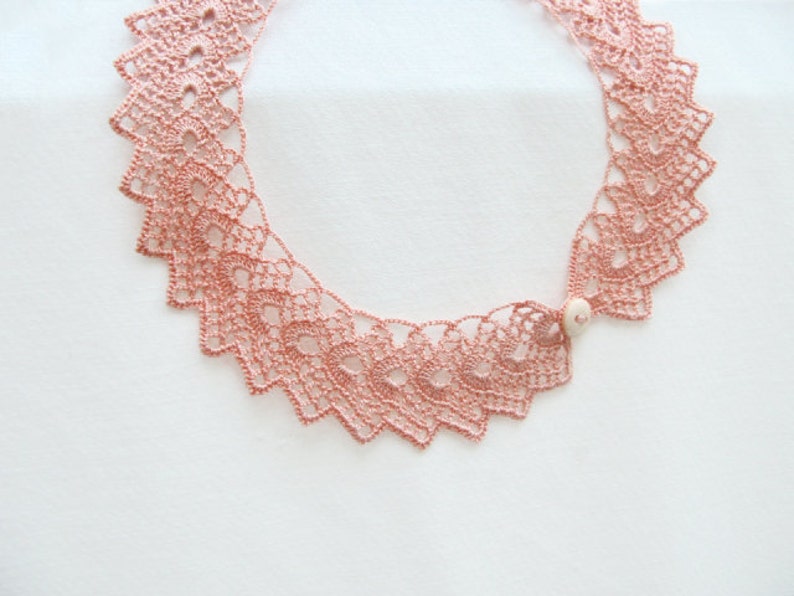 May include: A delicate pink crocheted necklace with a white bead accent. The necklace is made of intricate lacework and has a scalloped edge.