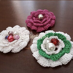 Peut inclure: Trois broches en forme de fleurs au crochet, faites à la main, en blanc, violet et vert. La fleur blanche a des perles rouges et nacrées, la fleur violette a une perle argentée, et la fleur verte et blanche a des perles nacrées et rouges. Les broches sont sur une surface marron.