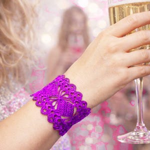May include: A vibrant purple crochet cuff bracelet with a floral design. The bracelet is worn on a wrist, and a champagne flute is held in the hand. The background is blurred with bokeh lights.