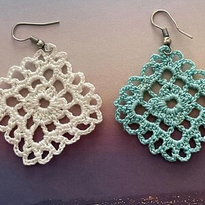 May include: A pair of crocheted earrings, one white and one light blue. The earrings are square shaped with a delicate floral design.