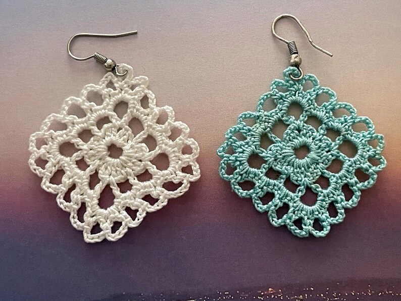 May include: A pair of crocheted earrings, one white and one light blue. The earrings are square shaped with a delicate floral design.