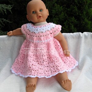 May include: A pink crocheted dress with white trim and a white crocheted collar. The dress is worn by a doll with blue eyes.
