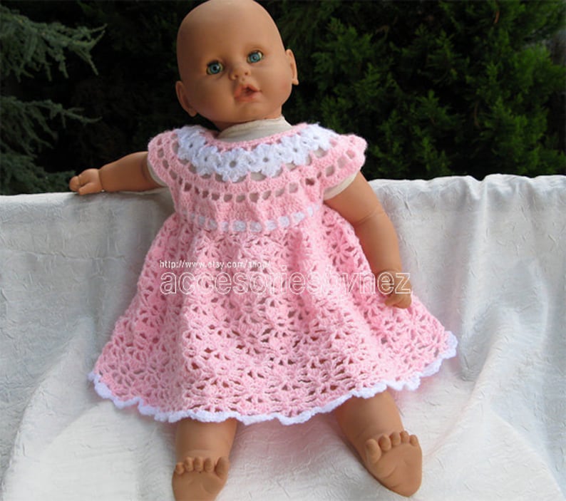 May include: A pink crocheted dress with white trim and a white crocheted collar. The dress is worn by a doll with blue eyes.