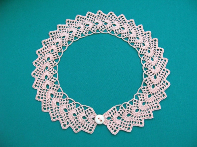 May include: A delicate pink crocheted collar with a white button closure. The collar is made of intricate lacework and has a scalloped edge.