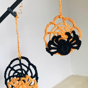 May include: Two crocheted spider web ornaments with spiders hanging from a black rod. One spider is black and the other is orange.