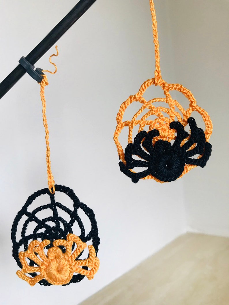 May include: Two crocheted spider web ornaments with spiders hanging from a black rod. One spider is black and the other is orange.