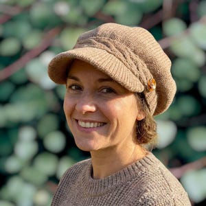May include: A tan knit hat with a small brim and a decorative cable knit design. The hat features a large, round, brown button on the side. The person in the image is wearing a matching tan sweater.