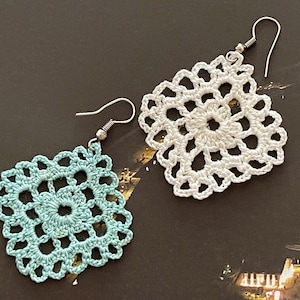 May include: A pair of crocheted earrings, one is white and the other is light blue. Both earrings are square shaped with a floral design.