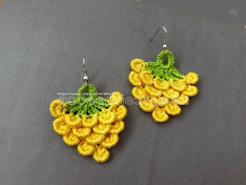 May include: A pair of crocheted earrings shaped like bunches of grapes. The earrings are yellow and green and have a silver hook.