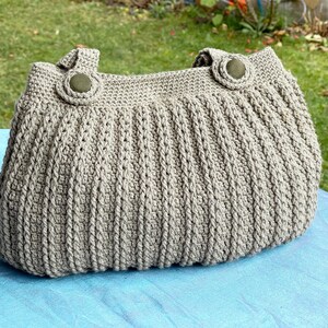 May include: A grey crocheted tote bag with two brown buttons. The bag is sitting on a blue surface.