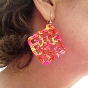 May include: A pair of crocheted earrings in a square shape with a geometric pattern. The earrings are made with pink, orange, and yellow yarn.