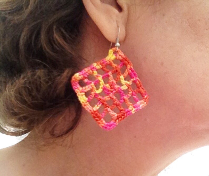 May include: A pair of crocheted earrings in a square shape with a geometric pattern. The earrings are made with pink, orange, and yellow yarn.