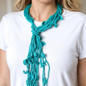 Puede incluir: Un collar turquesa de ganchillo con un diseño en cascada. El collar está hecho de un hilo suave y texturizado y presenta adornos en forma de hojas. El collar se usa sobre una camiseta blanca.
