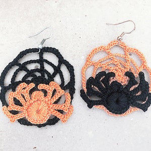 May include: A pair of crocheted spider earrings with black and orange details. The earrings are shaped like a spider web with a spider in the center.
