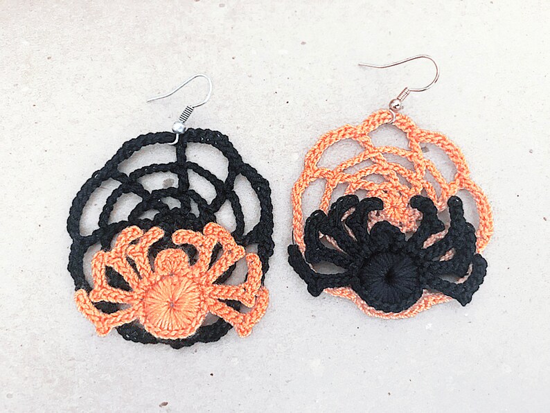 May include: A pair of crocheted spider earrings with black and orange details. The earrings are shaped like a spider web with a spider in the center.