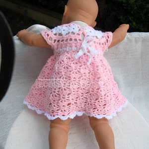 May include: A pink crocheted baby dress with white trim and a white bow on the back. The dress has a scalloped hem and a delicate crochet pattern.