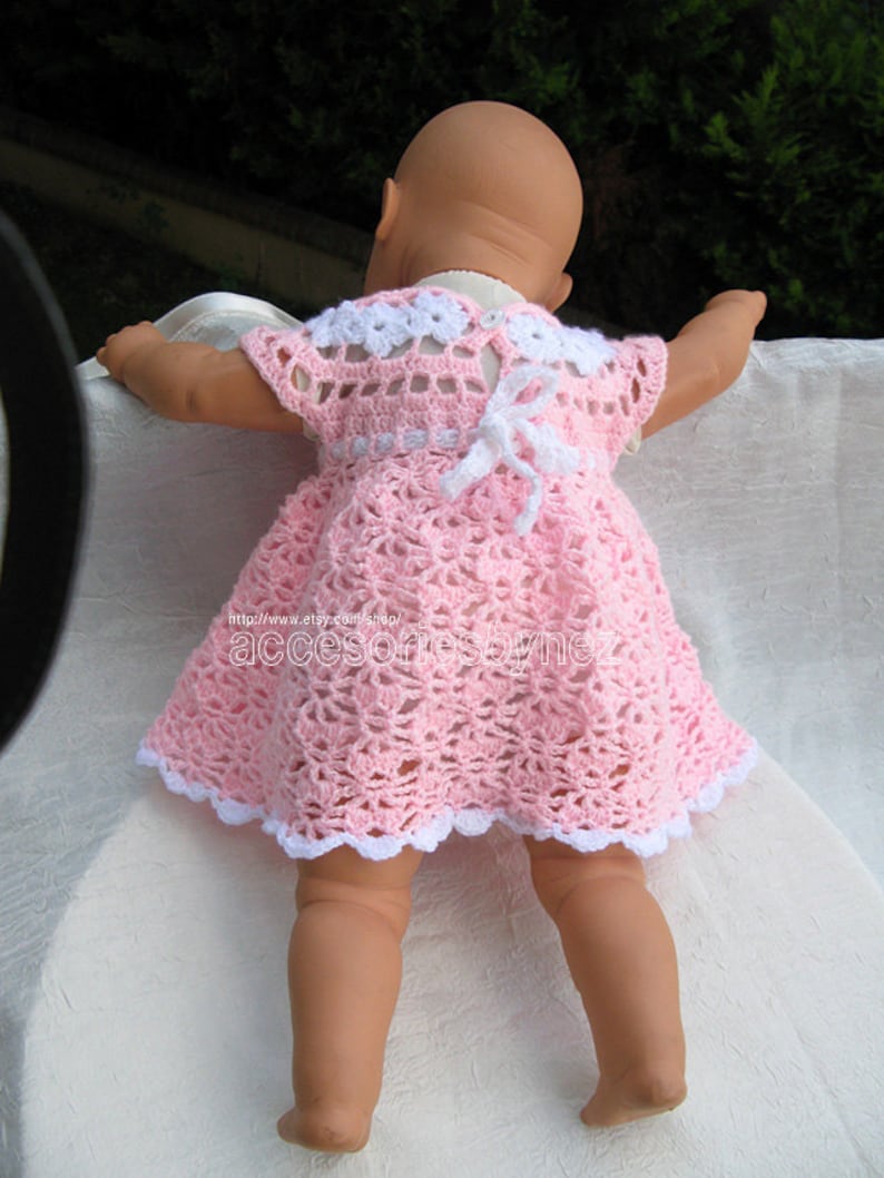 May include: A pink crocheted baby dress with white trim and a white bow on the back. The dress has a scalloped hem and a delicate crochet pattern.
