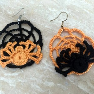 May include: A pair of crocheted spider earrings with black and orange webs. The earrings are made with a black web and a crocheted orange spider in the center.