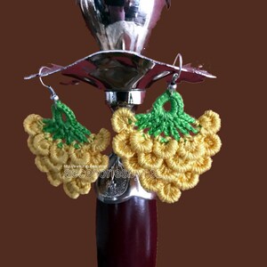 May include: A pair of crocheted grape earrings with yellow grapes and green leaves. The earrings are hanging from a silver hook.