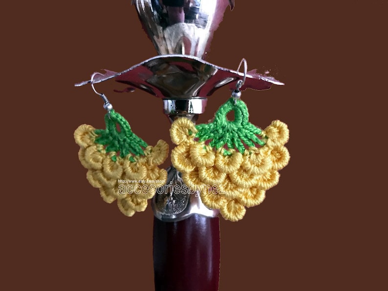 May include: A pair of crocheted grape earrings with yellow grapes and green leaves. The earrings are hanging from a silver hook.