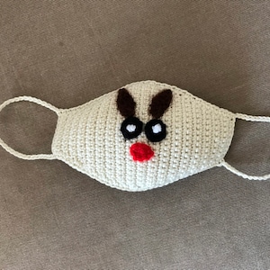 Puede incluir: M&aacute;scara facial de crochet blanca con un dise&ntilde;o de reno. El reno tiene cuernos marrones, ojos negros y una nariz roja.