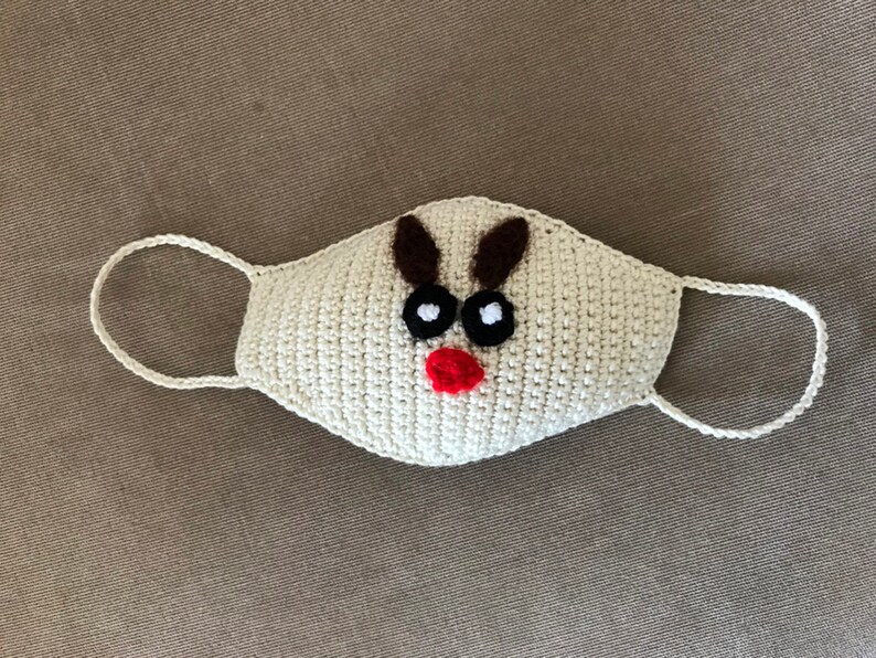 Puede incluir: M&aacute;scara facial de crochet blanca con un dise&ntilde;o de reno. El reno tiene cuernos marrones, ojos negros y una nariz roja.