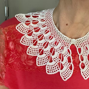 Puede incluir: Un collar blanco de ganchillo con un borde festoneado. El collar está hecho de encaje delicado y tiene un cierre de botón.