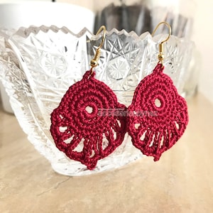 May include: A pair of crocheted earrings in a deep red color. The earrings are made with a delicate, lacy pattern and feature a small circle in the center. The earrings are attached to gold-colored hooks.