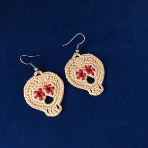 May include: A pair of crocheted skull earrings with red bead accents. The earrings are made with a light beige yarn and have silver hooks.