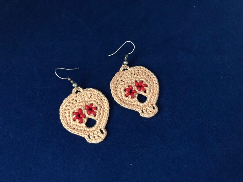May include: A pair of crocheted skull earrings with red bead accents. The earrings are made with a light beige yarn and have silver hooks.