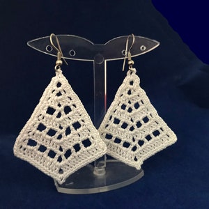 Peut inclure: Une paire de boucles d'oreilles en crochet blanc avec un motif géométrique. Les boucles d'oreilles sont suspendues à des crochets argentés sur un support en plastique transparent.