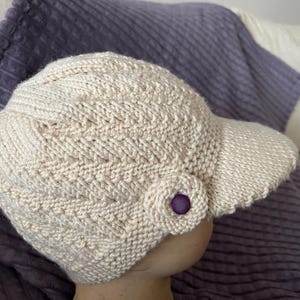 May include: A cream-coloured knitted hat with a visor. The hat features a textured pattern and a decorative purple button. The visor has a scalloped edge. The hat is shown against a purple background.
